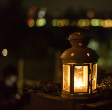 Lanterna decorativa acesa sobre muro com luzes desfocadas ao fundo à noite.