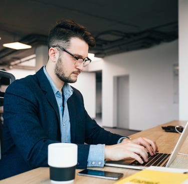 a man in a navy jacket and chambray shirt, sitting at a desk, working on a laptop computer