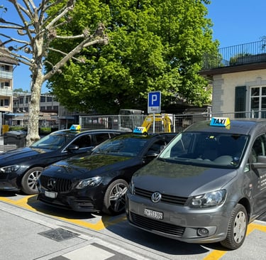Three taxicars. One grey VW Caddy and two black Mercedes E-Classes parked on a swiss mainstation.