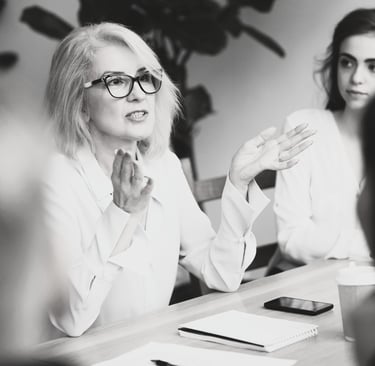 a woman in a white shirt and glasses sitting at a meeting table gesturing with her hands