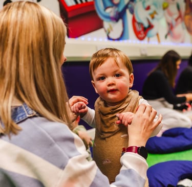 A young boy staring into the camera, holding his mums hands to stand up in moo class.