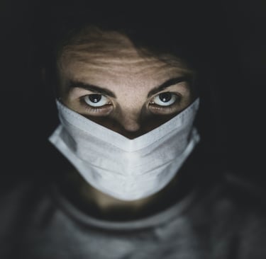Individual with a white face mask and visible eyes against a dark background, highlighting awareness