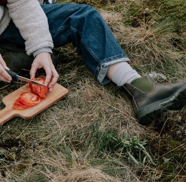 A pereson sitting on the grass cutting a tomato on a cutting board.