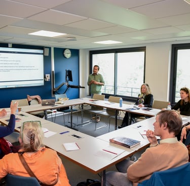 Un groupe d'adulte sont réunis autour d'une table carrée. Ils suivent la conférence d'un intervenant