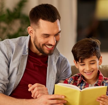 father and son are sitting on a couch reading a book