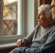 Ältere Frau sitzt am Fenster, hält die Hand eines Angehörigen, ruhige und liebevolle Szene