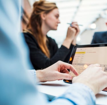 Person using a tablet during a meeting, with colleagues in discussion in the background.