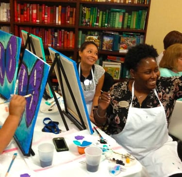 a smiling and laughing group of people sitting at a table painting on canvases