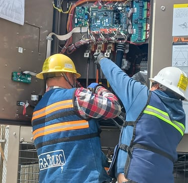 Dos técnicos con casco y chaleco trabajan en panel eléctrico abierto con circuitos.