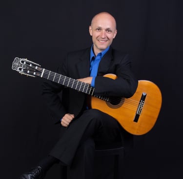 Antonio Gomez con ambo negro y camisa azul francia sentado con su guitarra en las manos
