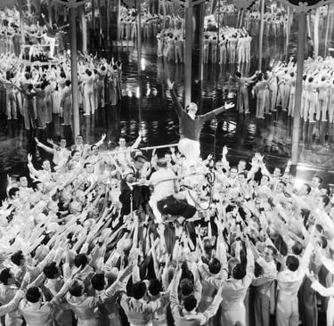 Busby Berkeley demonstrating arm movements to dancers on a mirrored stage platform.