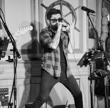 Singer Leo Couto performing on stage with a microphone, wearing a hat, black jeans, a plaid shirt, and sunglasses.