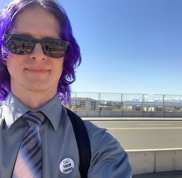 Tomkins with purple hair, wearing a gray shirt with a blue and pink tie, standing on a bridge in SLC