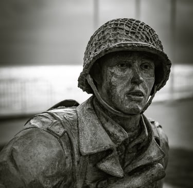 Gros plan sur le visage d'une statue de soldat en bronze, casque sur la tête, regard intense. Mer et ciel couvert en fond.