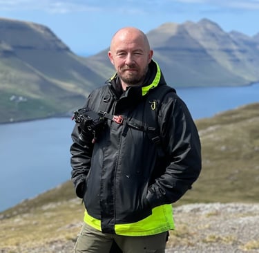 Bogi Olsen, your local Faroe Islands photography guide, standing outdoors with camera gear and scenic fjords behind him