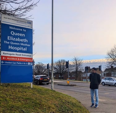 A man standing at a distance looking at the signpost reading 'Welcome to Queen Elizabeth The Queen Mother Hospital'