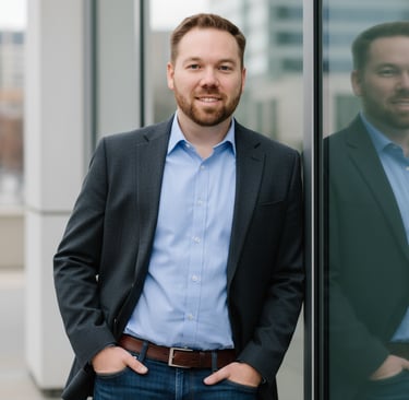 Man in blazer and jeans leaning against glass wall with city buildings in background.
