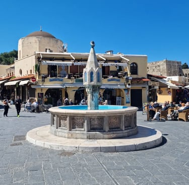 Historic Castellania Fountain in Rhodes Old Town square with cafes and a domed mosque background.