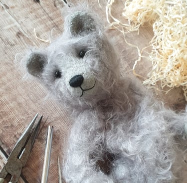a partly made handmade teddy bear in grey mohair with a wistful face and surrounded by tools on a table