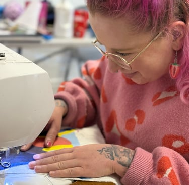 Harriet Mee, a woman with pink hair and glasses is sewing a picture