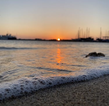 Foamy ocean waves wash onto a sandy beach at sunset with a harbor and boats in the background.