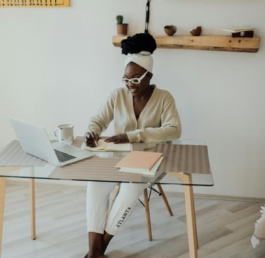 a woman sitting at a desk with an open laptop computer and writing in a notebook