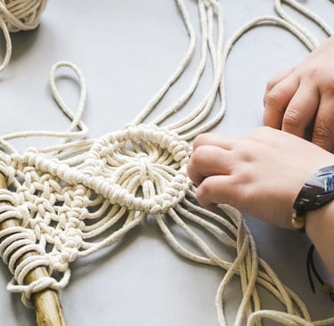 Manos elaborando un colgante de pared de macramé usando cordón de algodón blanco y una madera.