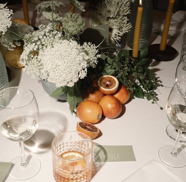 Elegantiškai dekoruotas stalas su baltomis gėlėmis, žalumynais, raudonais apelsinais ir kokteiliu