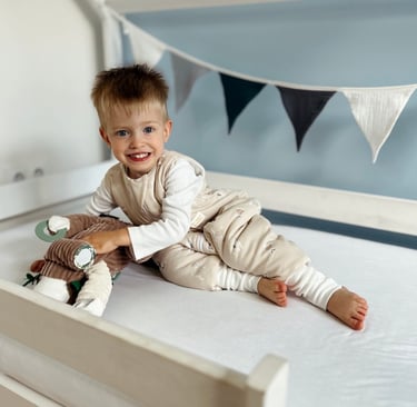 Uśmiechnięty chłopiec z jasnymi włosami w beżowym śpiworku z nogawkami leży na brzuchu w łóżeczku, t