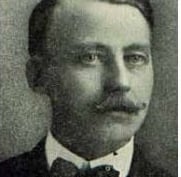 a headshot of a light eyed man with a curled mustache and a bowtie