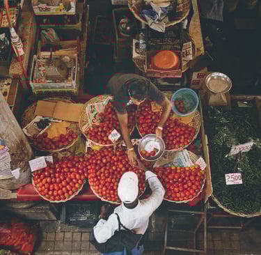 Overhead view of two men interacting at a market stall with baskets full of tomatoes, surrounded by various produce