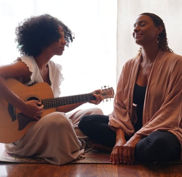 two women in front of a bright window sitting on a carpet playing a guitar.