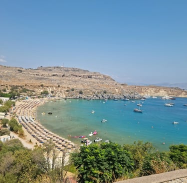 Panoramic view of Lindos beach in Rhodes with turquoise water, rows of umbrellas, and boats at anchor.