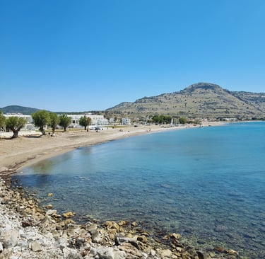 Panoramic view of a sandy Greek beach with turquoise sea, white buildings, and coastal mountains under a clear blue sky.