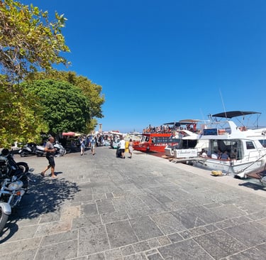 Crowded Mandraki Harbor in Rhodes Greece with tourist boats moored along the stone promenade under a blue sky.