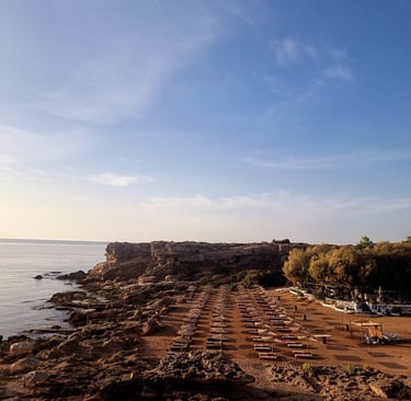 Seaside beach club with rows of sun loungers and umbrellas on a rocky coastline under a clear blue sky.