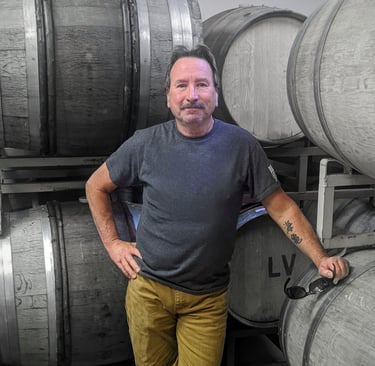 A man in a grey shirt leans against a wine barrel.