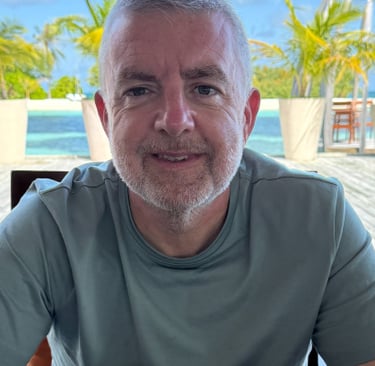 "Person seated at outdoor table in tropical resort with palm trees and lounge chairs nearby
