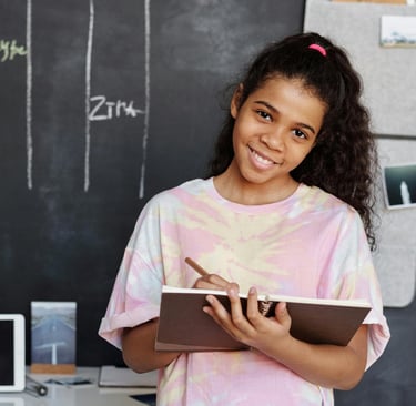 Menina adolescente sorridente segurando um caderno e lápis em frente a um quadro negro com cronograma semanal organizado