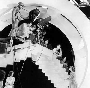 Busby Berkeley on a tall filming platform with cameras and lights, directing dancers below.