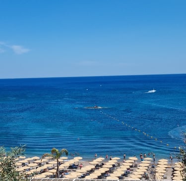 Wide view of a sunny beach resort with tan umbrellas and people swimming in deep blue ocean water.