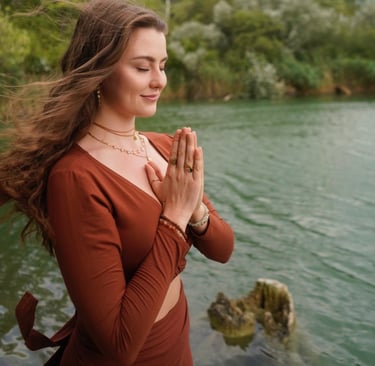 Celina Lerchner steht mit gefalteten Händen im Anjali Mudra vor einem See.