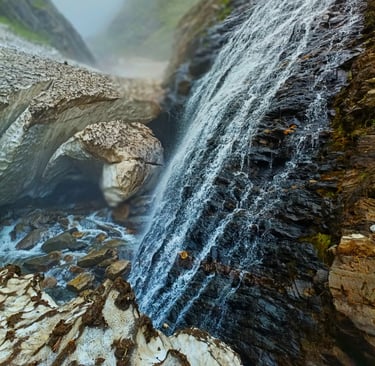 Snow-melt waterfall cascading over dark rocks in a misty mountain gorge