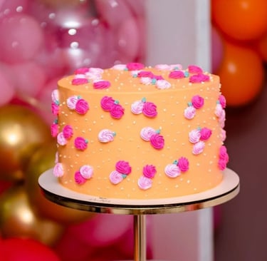 Bolo decorado com rosas coloridas em um suporte dourado, rodeado por balões em tons vibrantes. Ideal