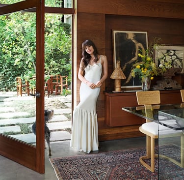 Dakota Johnson in white gown posing by glass doors at home.
