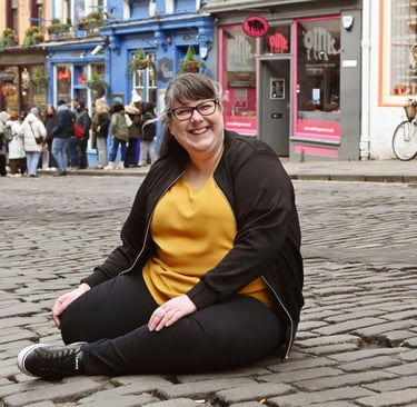 Laura is laughing at the camera sat on a cobbled street with her legs curled up under her