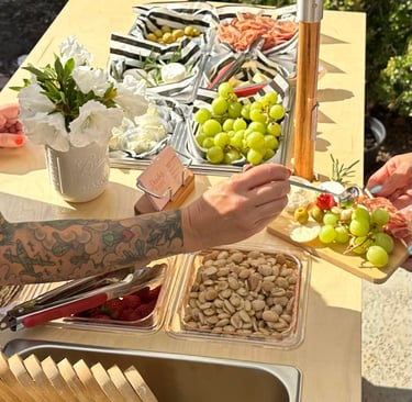 a person putting topping on a charcuterie board on a bar cart with charcuterie times in the backgrou
