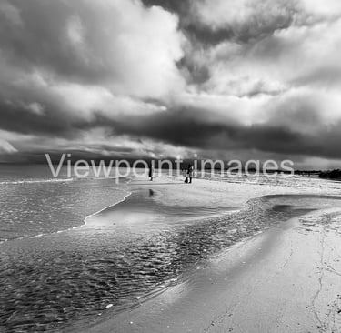 Dramatic black and white landscape of a sandy beach with dark storm clouds reflecting on the ocean tide.