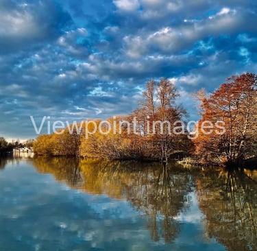 Tranquil river landscape with autumn trees reflecting in calm water under a dramatic blue sky.