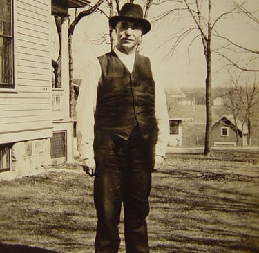 a gentleman in a vest and hat standing outside a house, black and white